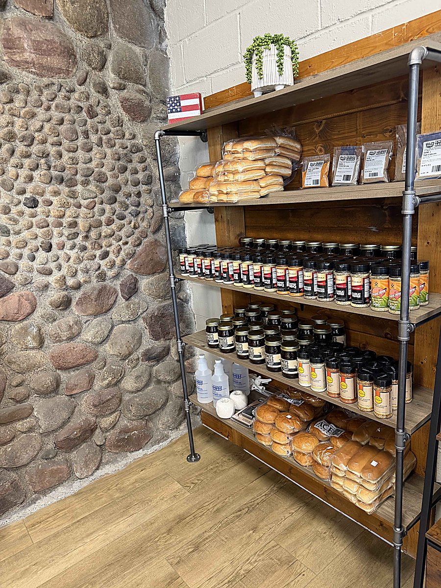 Industrial pipe shelving against a fieldstone wall stocked with BBQ sauces, rubs, hot sauces, honey, bread buns, and house favorites