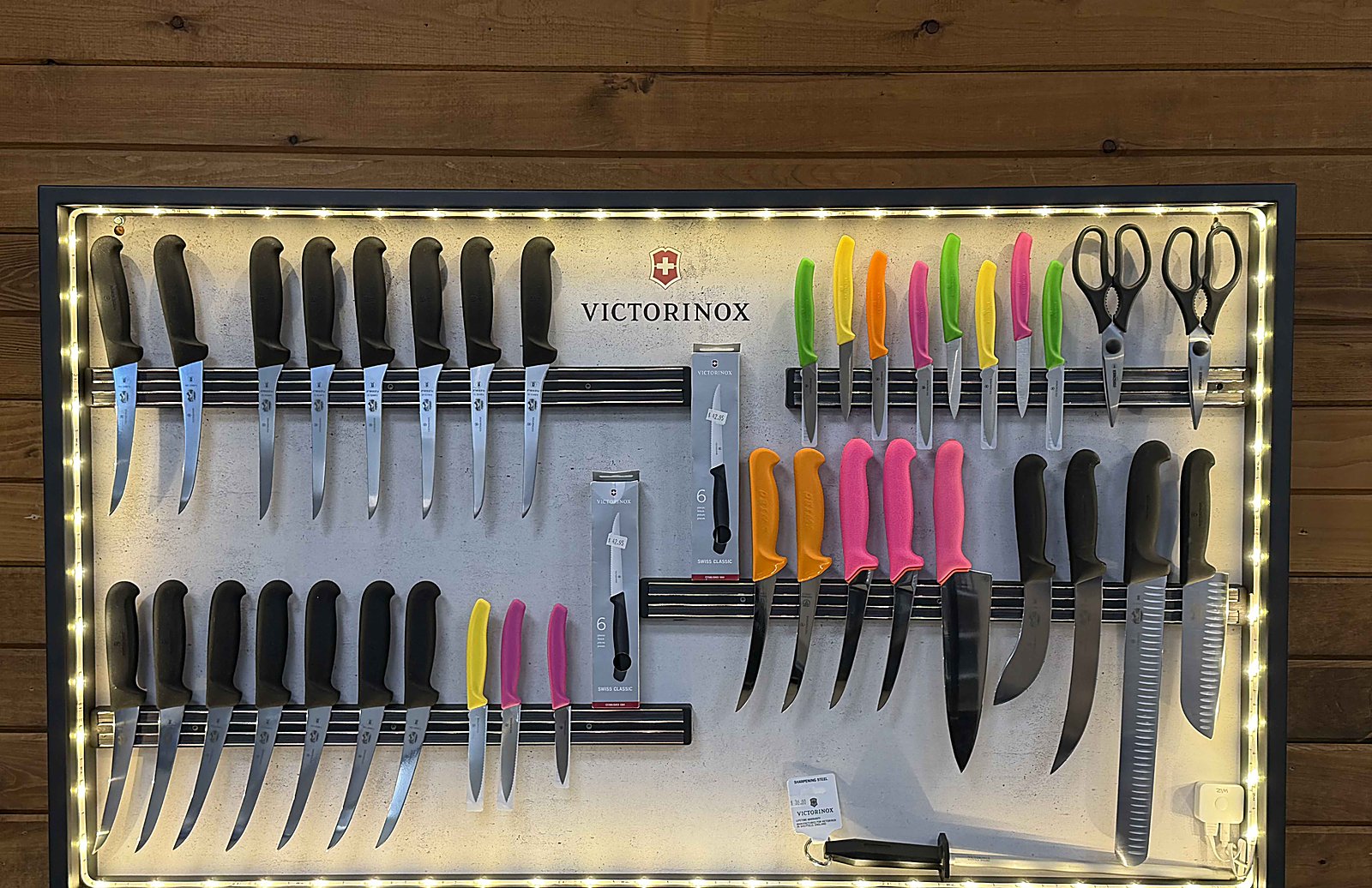 Backlit Victorinox retail display board with chef knives, santokus, paring knives with color-coded handles, kitchen shears, and sharpening steel