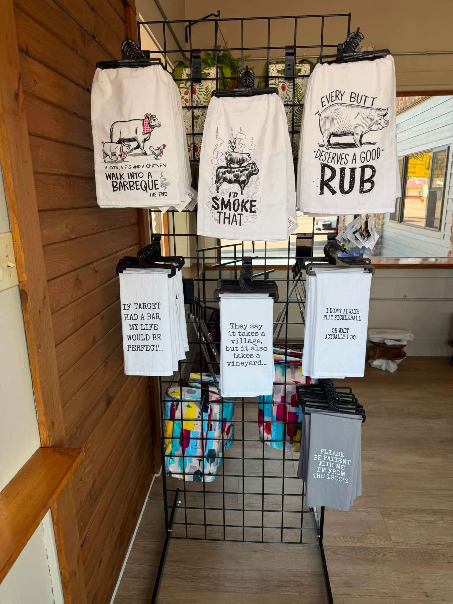 Rustic home goods display featuring humorous printed tea towels on a black wire rack, with a wood-paneled wall behind
