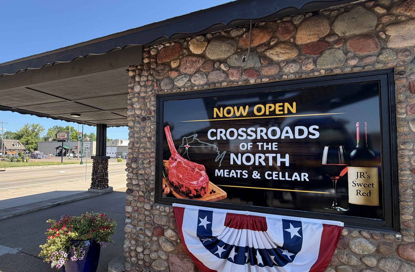 The Crossroads of the North Meats & Cellar storefront at the corner of Hwy 63 & Ash St., Spooner — stonework facade with the NOW OPEN sign and patriotic bunting